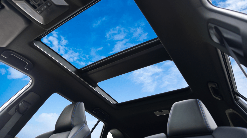 Looking up from the rear seats of a modern car interior, focusing on a large dual-panel panoramic sunroof revealing a deep blue sky.