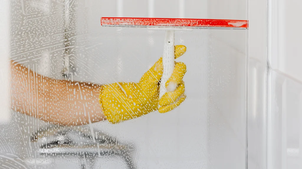 A shower glass door in the middle of being cleaned with a squeegee and gloves, highlighting best practices for cleaning shower glass.